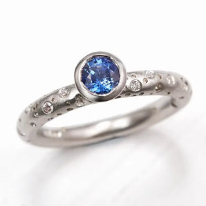 A white gold engagement ring featuring a central blue sapphire with patterns and brilliant cut diamonds around it.