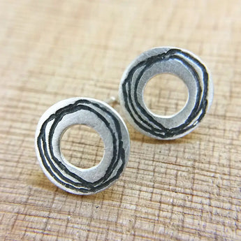 A pair of circular silver earrings with spiral etched details on a satin finish, displayed against a wooden background.