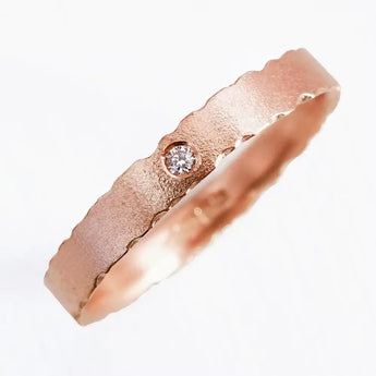 A rose gold engagement ring with a central diamond gemstone, featuring subtly burnished edges and a slightly eroded texture.