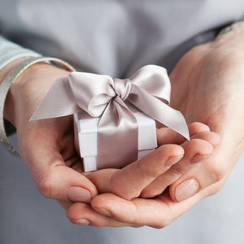 Small gift box with a pink ribbon held in hands against a neutral background Kate Smith Jewellery Birmingham
