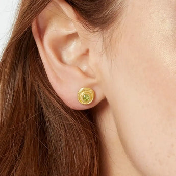 A close-up image of a woman's ear wearing a gold stud earring with a green peridot in the center.