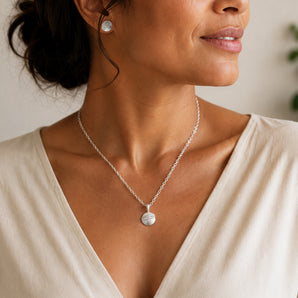 A close-up lifestyle shot of a woman wearing a delicate silver chain necklace with a textured circular pendant. She is wearing a cream-colored V-neck top, and the background is a soft, out-of-focus neutral wall.