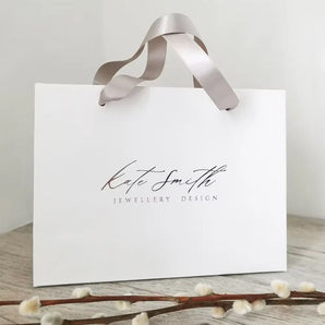 Lifestyle shot of a branded Kate Smith Jewellery gift bag in white with taupe satin ribbon handles.