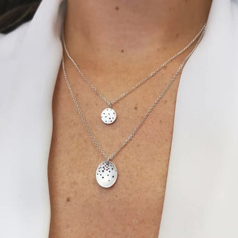 A person wearing a white shirt layering two silver pendant necklaces, one round and one oval, with a background of a light-colored fabric.