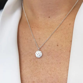 A person wearing a silver pendant necklace with a round shape and patterns resembling seed heads, on a white shirt.