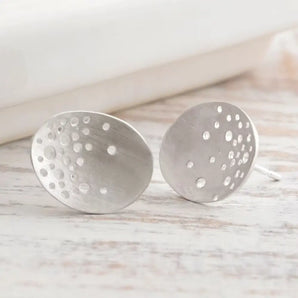 A pair of silver oval earrings with hand drilled patterning, displayed against a wooden background.
