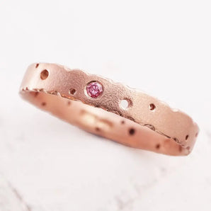 A rose gold ring with a central pink diamond surrounded by a pattern of nibbled marks on a white background.