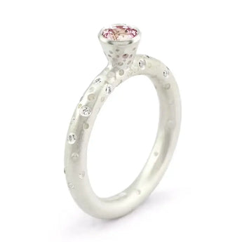 A silver engagement ring featuring a central pink sapphire with brilliant cut diamonds on the sides, with a patterned band.