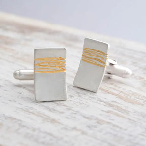A pair of silver cufflinks with gold etched detail, placed on a wooden surface.