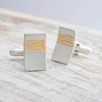 A pair of silver cufflinks with gold etched detail, placed on a wooden surface.