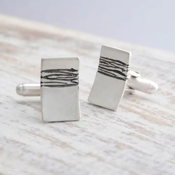 A pair of silver cufflinks with etched details and oxidised blackening, displayed on a wooden surface.