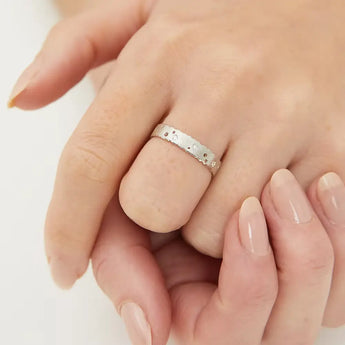 A silver diamond eternity ring with patterned design and scattered diamonds, displayed on a person's hand.