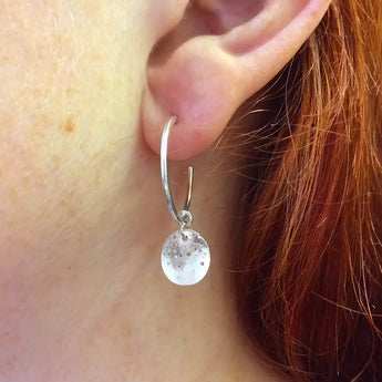 A close-up image of a person wearing silver hoop earrings with a patterned disc detail.