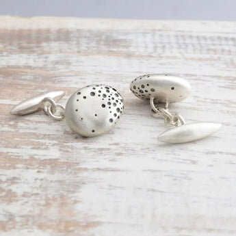 A pair of handmade sterling silver nugget cufflinks with chain fittings and blackened dots on a wooden surface.