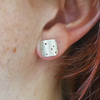 A close-up image of a person wearing a silver square textured earring with hand-drilled patterning.