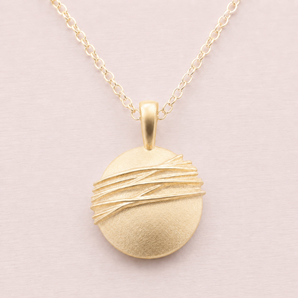Plain pale background with close up of a round handmade in Birmingham gold pendant and fine chain. The disc has raised irregular diagonal lines which are polished on a matt satin background.
