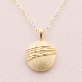 A gold pendant necklace featuring a textured, matte circular disc pendant wrapped with three parallel metallic wire strands, suspended from a delicate gold chain against a soft, light pink background.