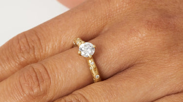 Gold ring with a diamond on a person's finger against a neutral background