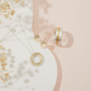 Gold jewelry including a necklace, earrings, and rings on a soft pink background with flowers.