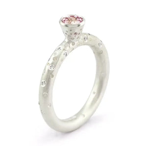 A silver engagement ring featuring a central pink sapphire with brilliant cut diamonds on the sides, with a patterned band.