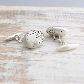 A pair of handmade sterling silver nugget cufflinks with chain fittings and blackened dots on a wooden surface.