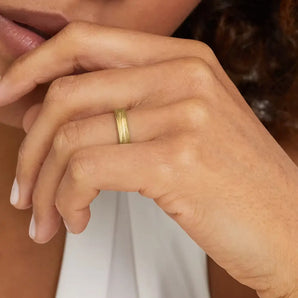 A close-up image of a person's hand wearing a slim gold wedding ring with texture reminiscent of windswept sand dunes.