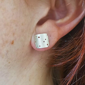 A close-up image of a person wearing a silver square textured earring with hand-drilled patterning.